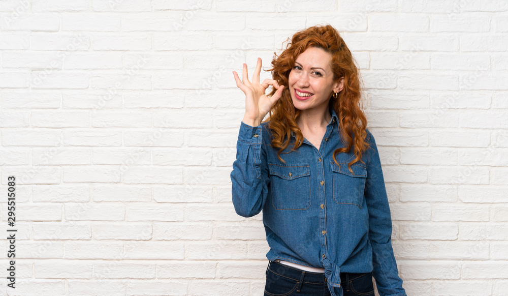 Redhead woman over white brick wall showing ok sign with fingers Redhead woman over white brick wall showing ok sign with fingers