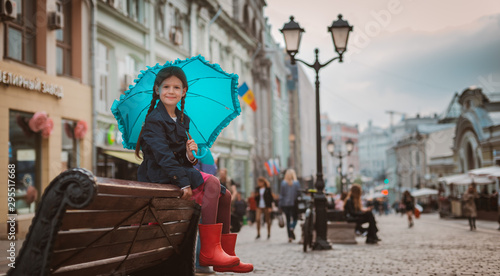 Little girl 6 years old child with an umbrella in rubber boots having fun on a bench in the center of Moscow in the fall or spring.
