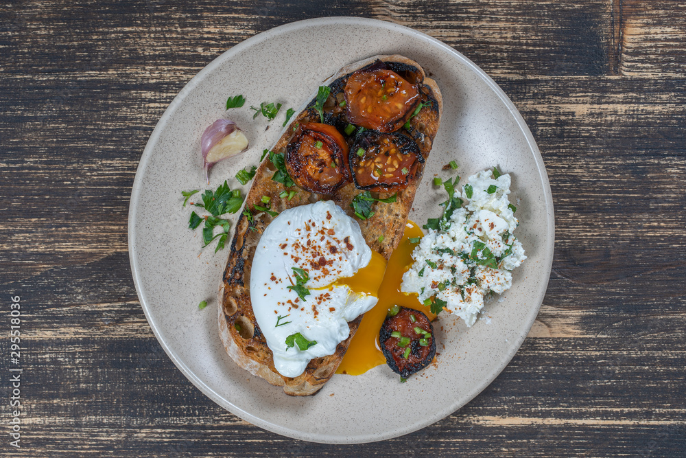 Poached egg on a piece of bread with fried red tomato, garlic and cottage cheese on a plate, close up