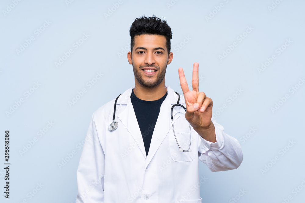 Young doctor man over isolated blue wall smiling and showing victory sign Young doctor man over isolated blue wall smiling and showing victory sign