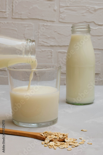 Oat milk pouring into a glass cup with a bottle of milk on the background, selective focus