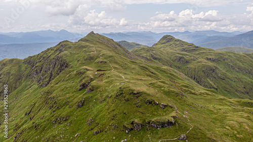 An aerial view of a grassy mountain rocky summit and mountain range in the background under a cloudy sky