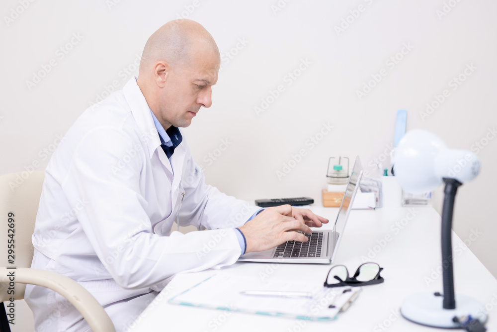 Serious middle aged doctor in whitecoat concentrating on laptop work