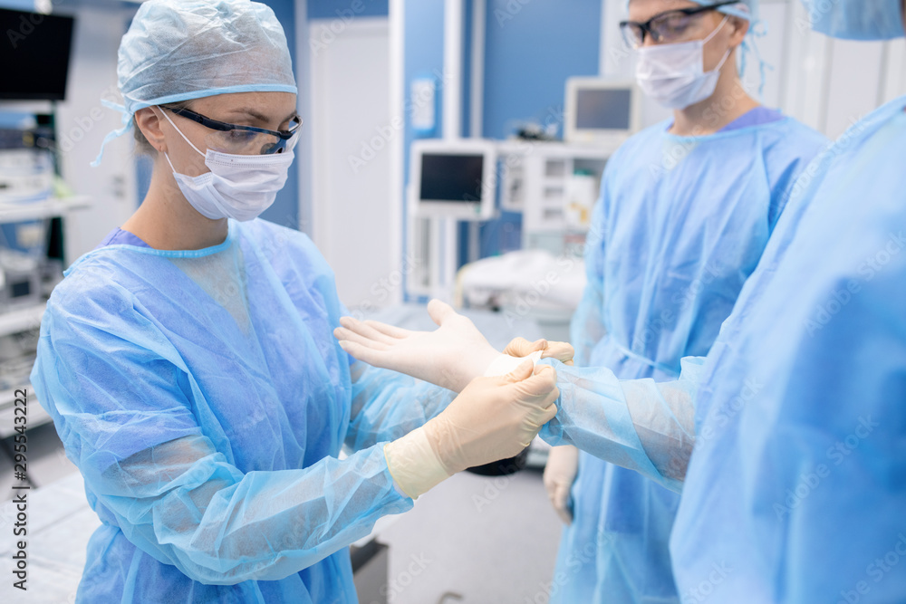 Young nurse in protective mask and workwear helping surgeon with gloves Young nurse in protective mask and workwear helping surgeon with gloves