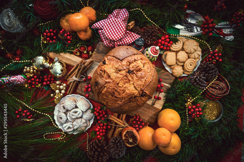 Christmas ingredients table with a Panettone in the center on a wooden board, accompanied by orange, cookies, cinnamon, pine. Festive pine decoration on red rustic background, top view