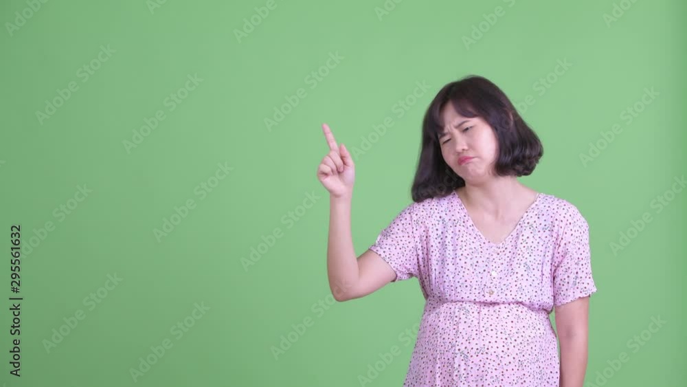 Stressed Asian pregnant woman pointing up and giving thumbs down Stressed Asian pregnant woman pointing up and giving thumbs down