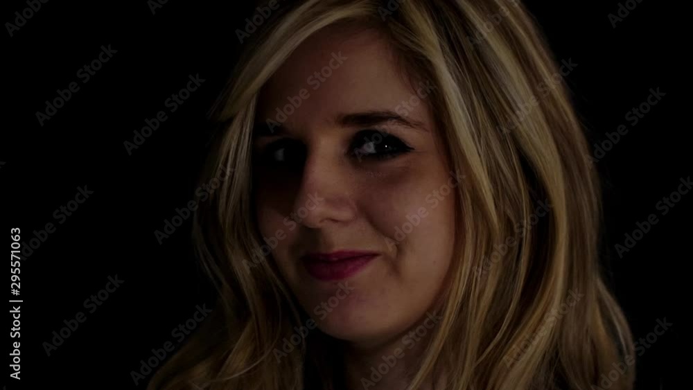 Beautiful blond woman smiling flirtatiously charmingly to camera; low key light. Beautiful blond woman smiling flirtatiously charmingly to camera; low key light.
