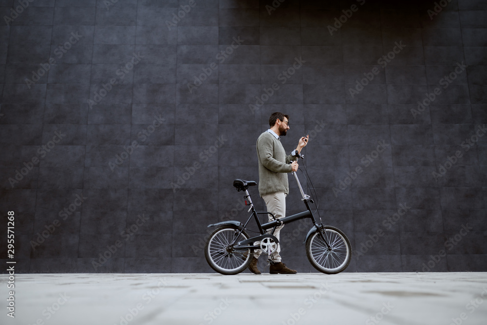 Full length of stylish caucasian man pushing bicycle and using smart phone while passing by gray wall. Full length of stylish caucasian man pushing bicycle and using smart phone while passing by gray wall.