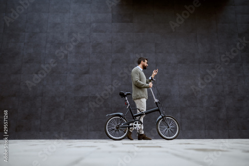 Full length of stylish caucasian man pushing bicycle and using smart phone while passing by gray wall.