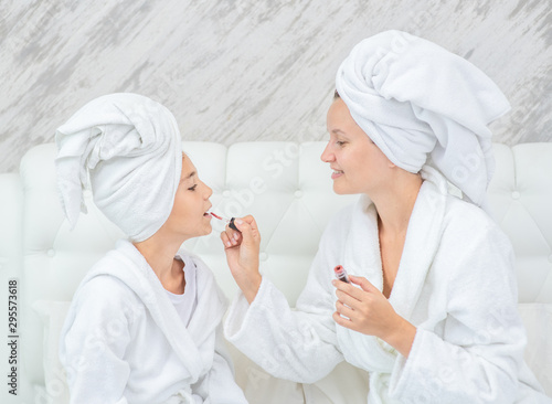 Happy family at home. Mom paints her daughter's lips on the bed at home. Mom and child girl are in bathrobes and with towels on their heads