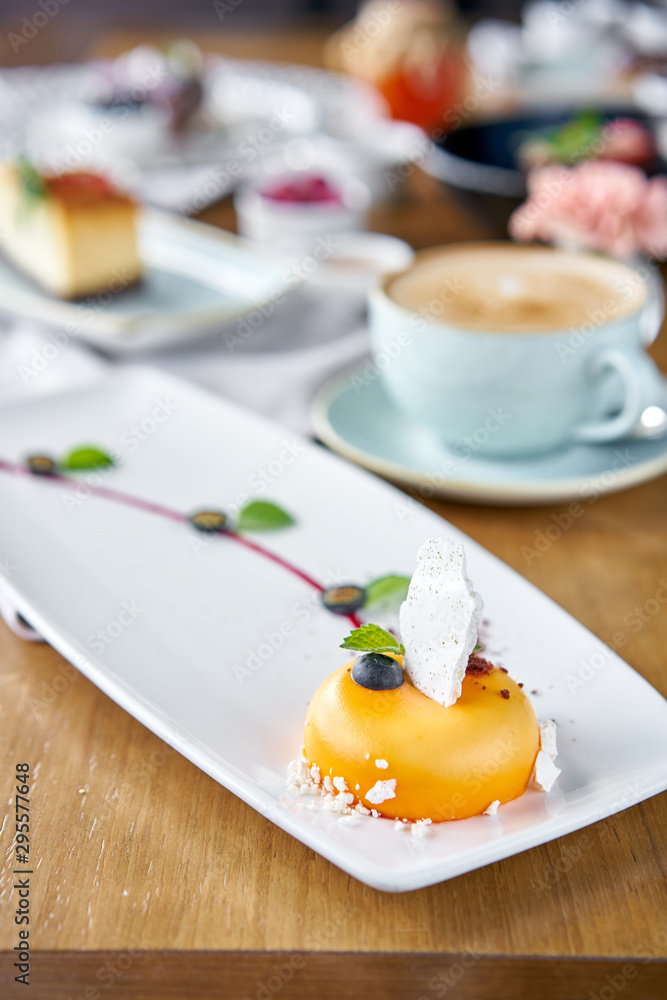 Yellow mousse-cake. Breakfast in the cafe, morning coffee. Cappuccino and lots of desserts on the table. French dessert, blueberry, mint and a piece of meringue on a white plate. Restaurant menu