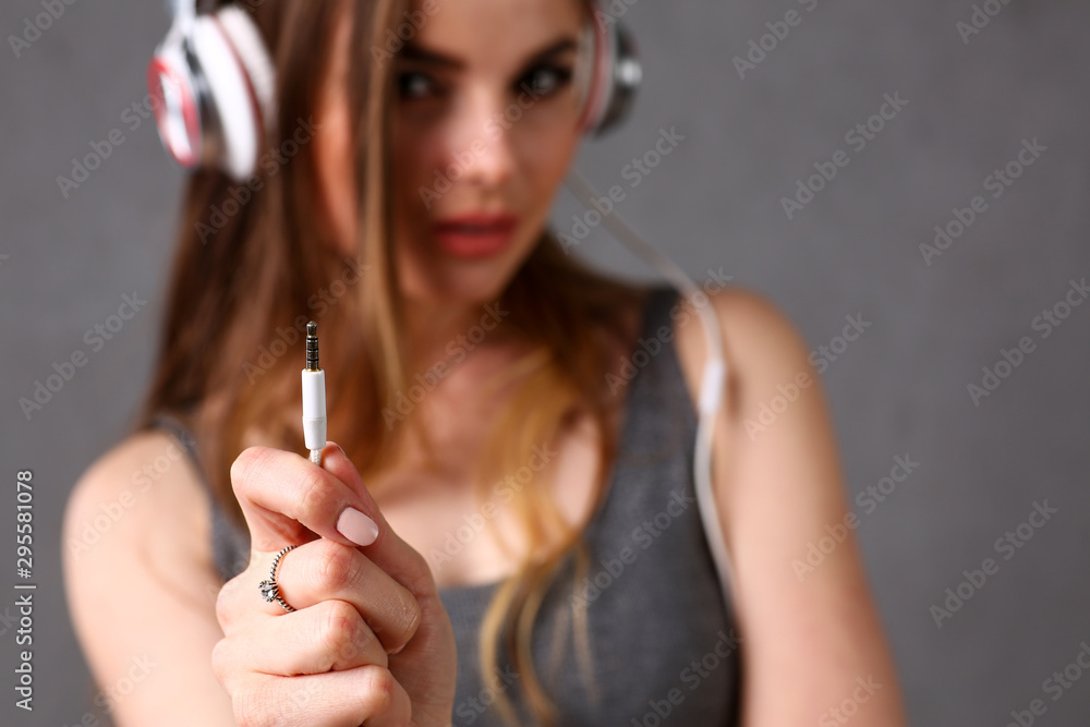Obraz premium Beautiful blonde woman wearing headphones listening favourite music hold cable jack in arm closeup on grey background. Modern urban life audio book radio having moments with dance move concept
