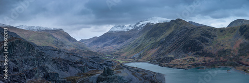 Beautiful landscape image of Dinorwig Slate Mine and snowcapped Snowdon mountain in background during Winter in Snowdonia with Llyn Peris in foreground