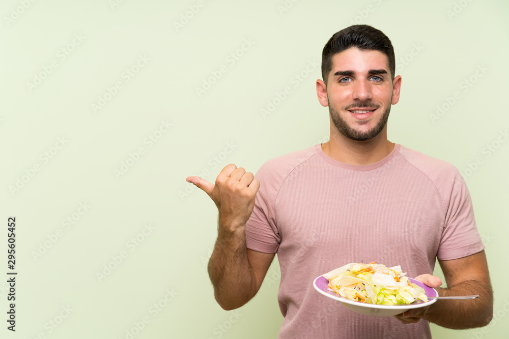 Young handsome man with salad over isolated green wall pointing to the side to present a product Young handsome man with salad over isolated green wall pointing to the side to present a product