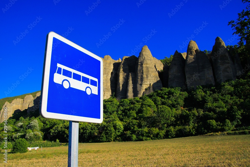 View on isolated bus stop sign against rural field, trees, needle like sharp bizarr rock formations (Les Pénitents) and blue sky - Les Mées, Provence, France