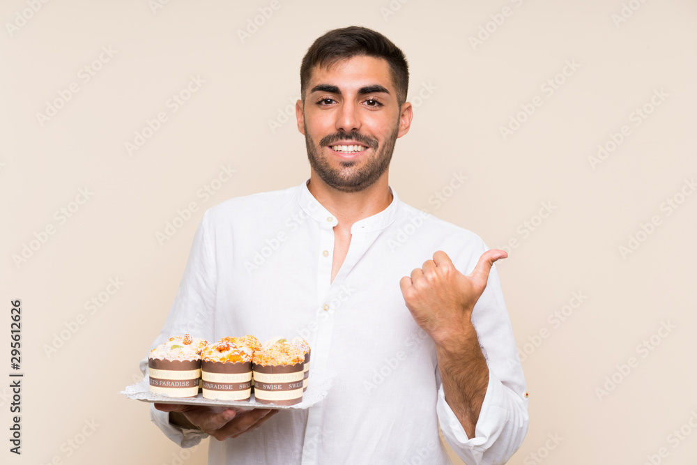 Handsome man holding muffin cake over isolated background pointing to the side to present a product Handsome man holding muffin cake over isolated background pointing to the side to present a product