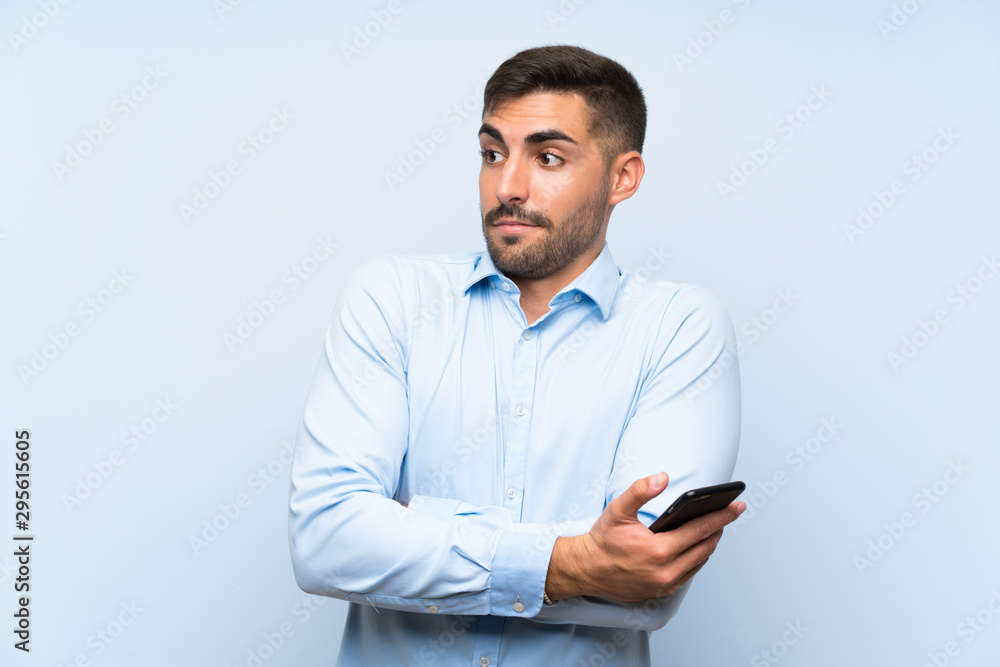 Young handsome man with his mobile over isolated blue wall making doubts gesture while lifting the shoulders Young handsome man with his mobile over isolated blue wall making doubts gesture while lifting the shoulders