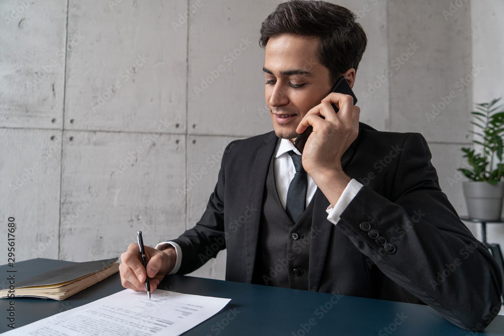Friendly young businessman with dark hair talking on smartphone and signing documents at loft office table. Concept of corporate lifestyle and management Friendly young businessman with dark hair talking on smartphone and signing documents at loft office table. Concept of corporate lifestyle and management