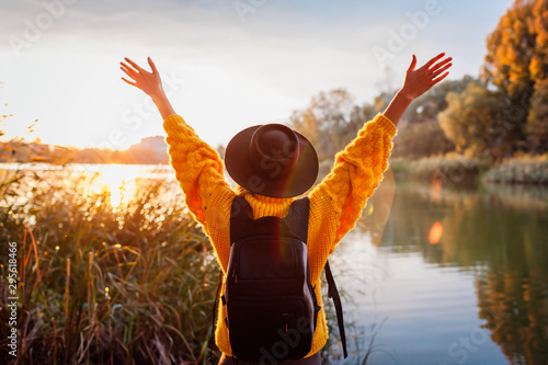 Traveler with backpack relaxing by autumn river at sunset. Young woman raised arms feeling free and happy