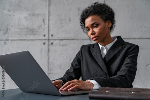 Side view of calm young African woman in business suit typing on laptop keyboard in blurred loft office. Concept of management and business planning