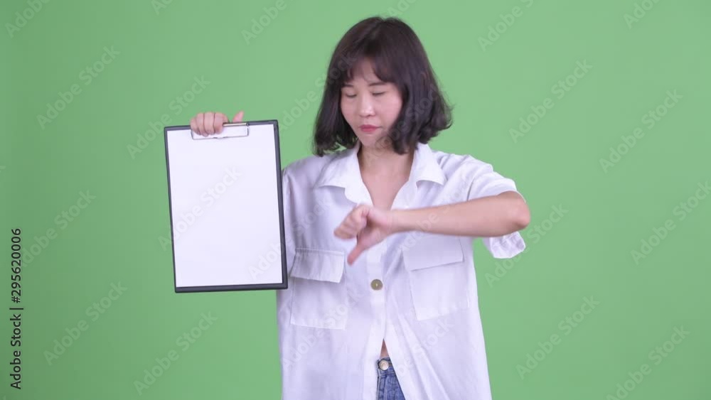 Stressed Asian businesswoman showing clipboard and giving thumbs down Stressed Asian businesswoman showing clipboard and giving thumbs down