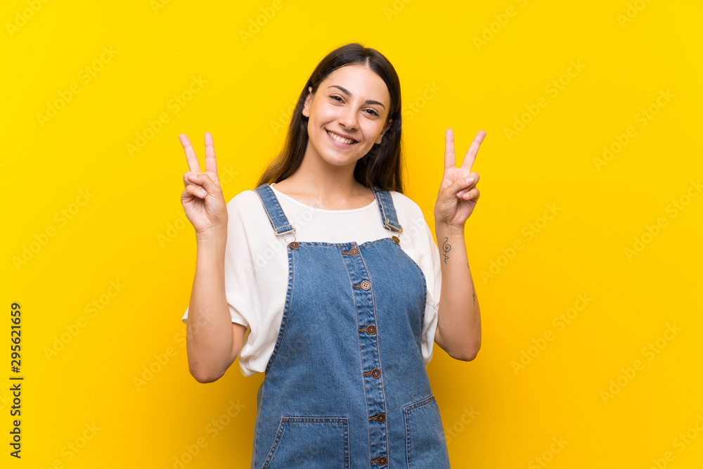 Young woman in dungarees over isolated yellow background showing victory sign with both hands Young woman in dungarees over isolated yellow background showing victory sign with both hands