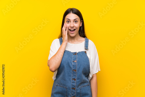Young woman in dungarees over isolated yellow background with surprise and shocked facial expression
