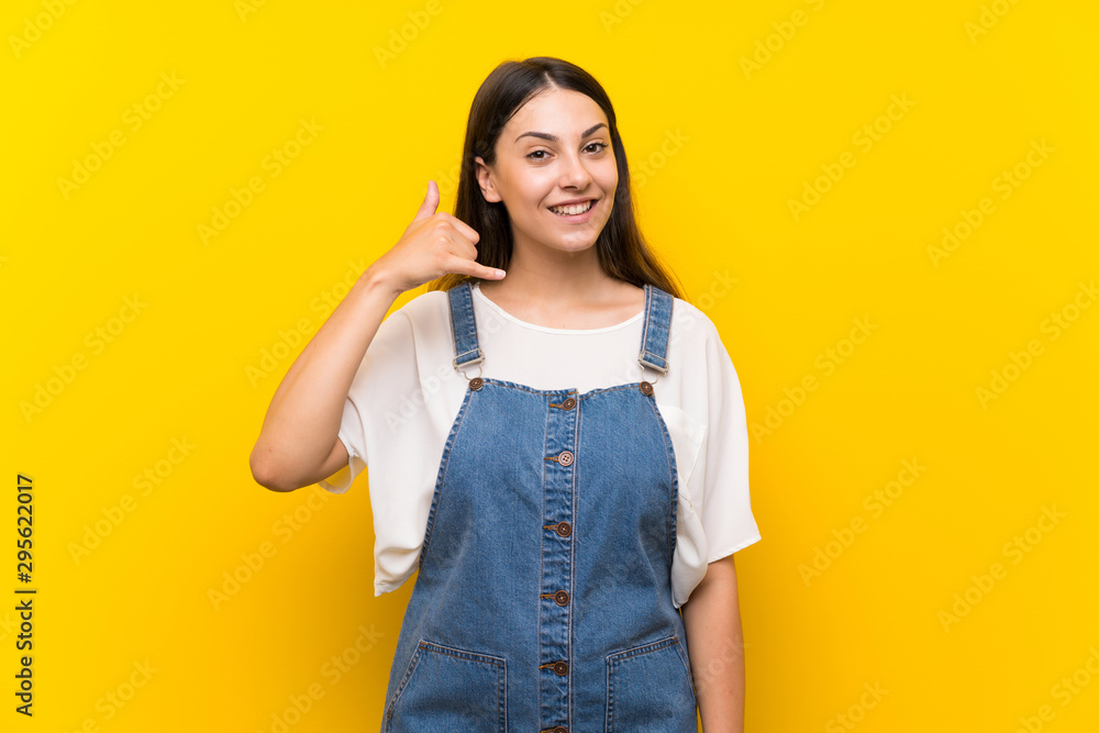 Young woman in dungarees over isolated yellow background making phone gesture. Call me back sign Young woman in dungarees over isolated yellow background making phone gesture. Call me back sign