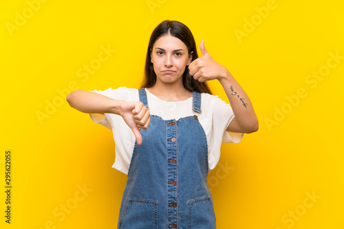 Young woman in dungarees over isolated yellow background making good-bad sign. Undecided between yes or not