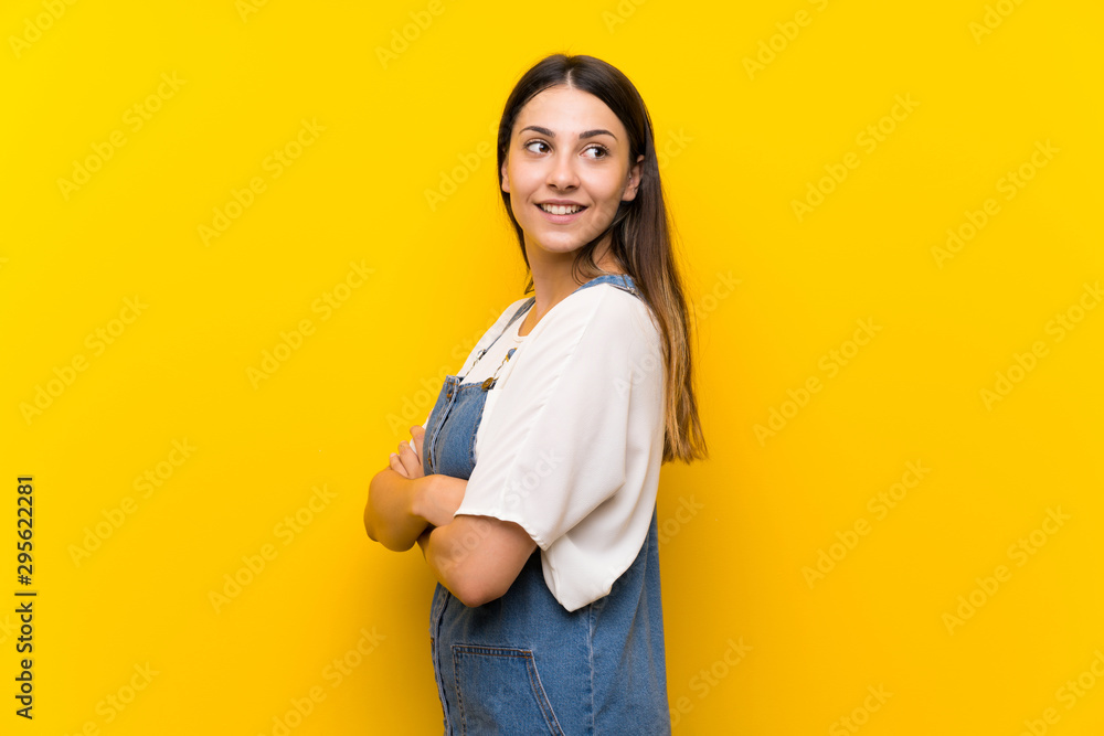 Young woman in dungarees over isolated yellow background with arms crossed and happy Young woman in dungarees over isolated yellow background with arms crossed and happy