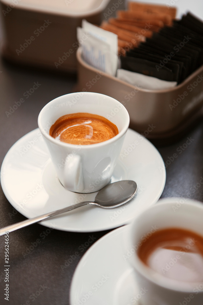 Close up. The small white ceramic mug (cup) with Italian espresso (coffee) on a table with a saucer and a metal spoon in a local Italian bar (coffee shop) in Milan, Lombardy, Italy. Beverage. Close up. The small white ceramic mug (cup) with Italian espresso (coffee) on a table with a saucer and a metal spoon in a local Italian bar (coffee shop) in Milan, Lombardy, Italy. Beverage.