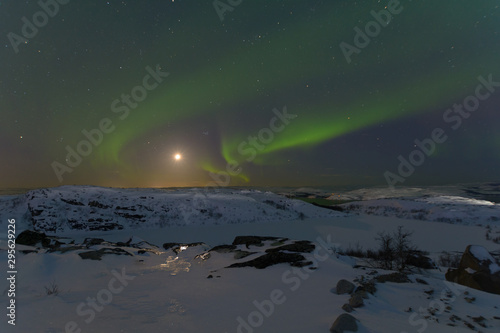 Northern lights  aurora in the sky at night. Hills and rocks covered with snow. Moon in the sky.