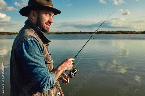 Caucasian adult bearded men stand near lake and hold fishing rod. Here he with his son, he shows how hold fishing rod correctly.