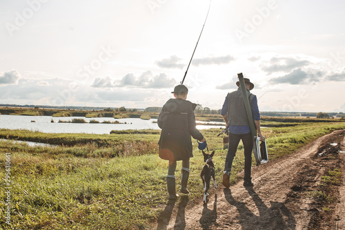 Father shows fisherman family trail to young son, who take dog and follow. Father and son carriy fishing gear together.