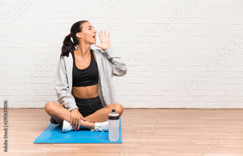 Young sport woman sitting on the floor with mat shouting with mouth wide open