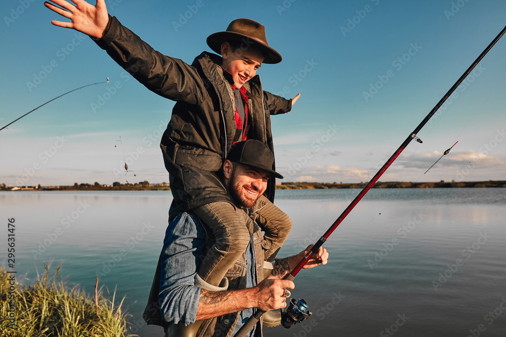 Son and father have fun while fishing. Father holds son on shoulders. Boy holds hands to sides. They smile. Background lake.