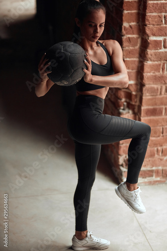 Flexible and beautiful sportswoman stretching legs with help of balls. Caucasian Girl in black sportswears. Brickwall background