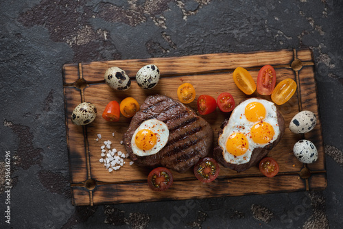 Grilled marbled beefsteaks with fried quail eggs and cherry tomatoes on a rustic wooden serving board, top view on a brown stone surface