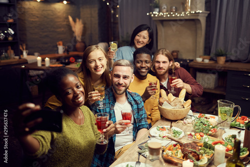 Multi-ethnic group of young people taking selfie photo via smartphone while enjoying dinner party at home, copy space
