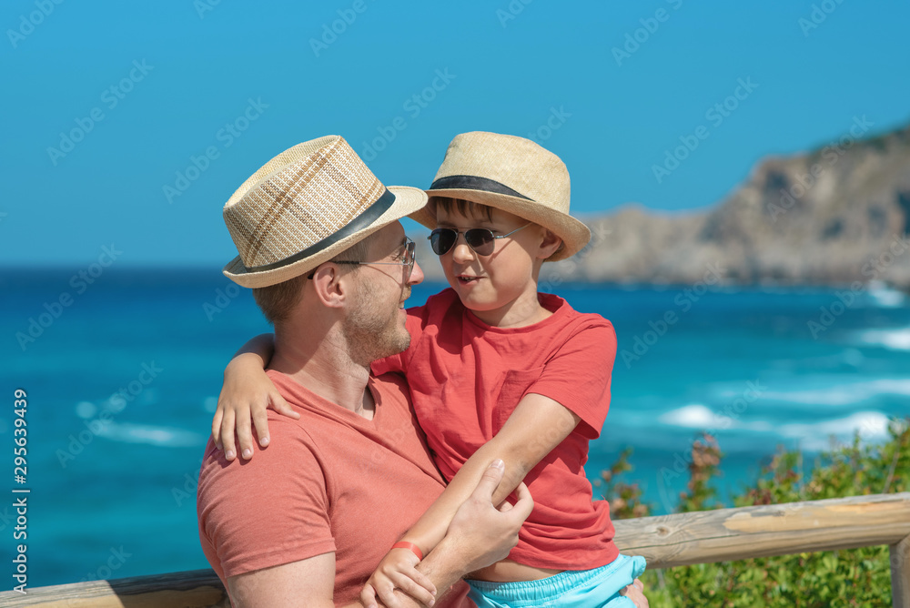 Cute European boy is on his father’s hands in front of picturesque wavy sea, enjoying summer holidays. They wearing similar clothes sun hats and sunglasses smiling and looking to each other.