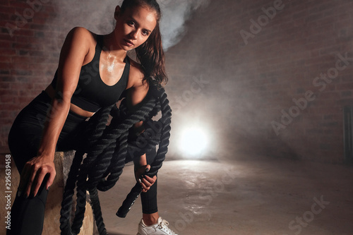 Young sporty female sit on wooden fit box in gym holding battle rope. Crossfit, sport, healthy lifestyle concept