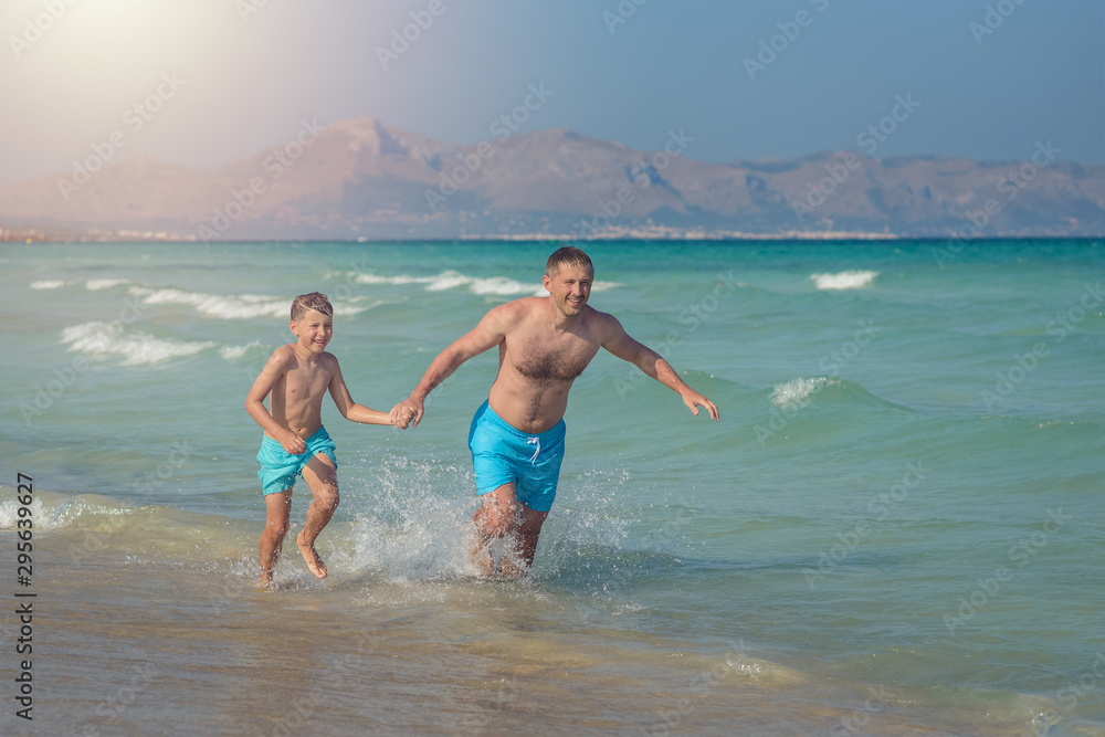 Father and son enjoying their summer holidays, they are running into the sea holding hands.