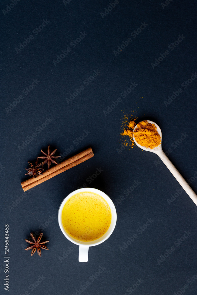 Golden (turmeric) milk in a white cup on the dark background. A spoon with turmeric powder. Cinnamon and anise. Golden (turmeric) milk in a white cup on the dark background. A spoon with turmeric powder. Cinnamon and anise.