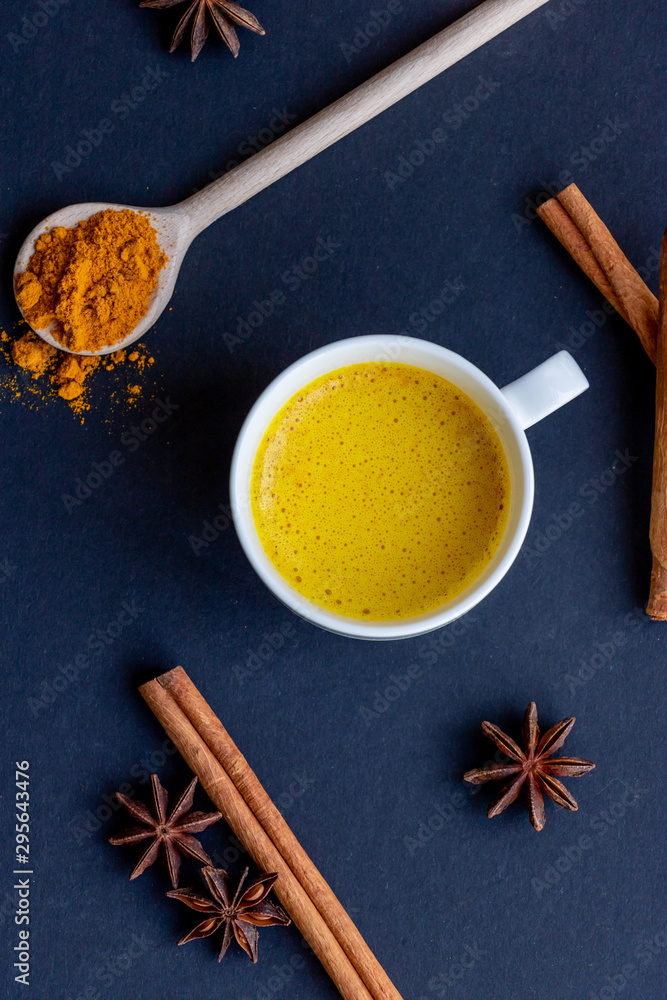 Golden (turmeric) milk in a white cup on the dark background. A spoon with turmeric powder.  Cinnamon and anise.