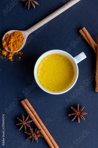 Golden (turmeric) milk in a white cup on the dark background. A spoon with turmeric powder. Cinnamon and anise.