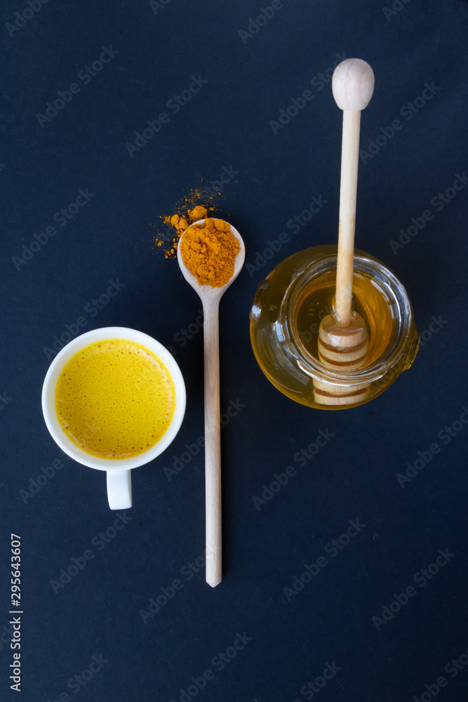 Golden (turmeric) milk in a white cup on the dark background. Jar with honey. A spoon with turmeric powder. Top view. Golden (turmeric) milk in a white cup on the dark background. Jar with honey. A spoon with turmeric powder. Top view.