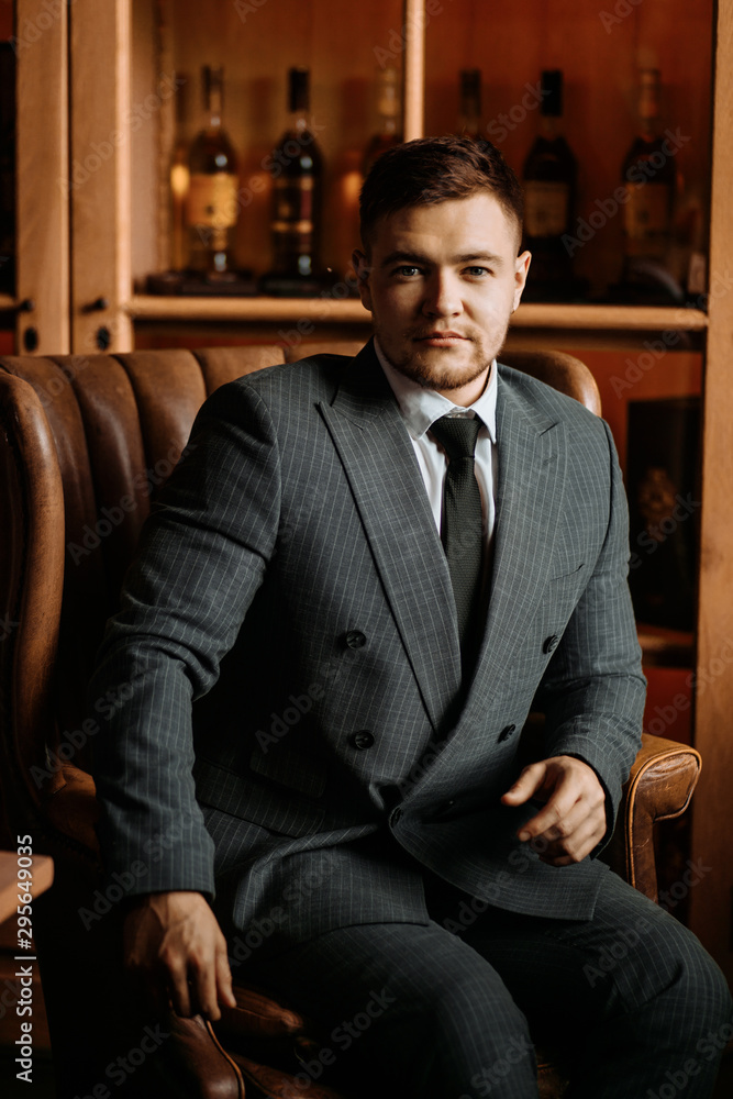 man in formal buttoned suit with a tie, sits respectfully on large leather armchair, puts hands on armrest, looks straight towards camera with courageous expression, posing over wall of bottles man in formal buttoned suit with a tie, sits respectfully on large leather armchair, puts hands on armrest, looks straight towards camera with courageous expression, posing over wall of bottles
