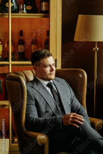 Portrait of confident bright businessman sitting thoughtfully in large soft armchair with crossed legs, puting right hand on knee, looking unemotion towards window, right side shot photo