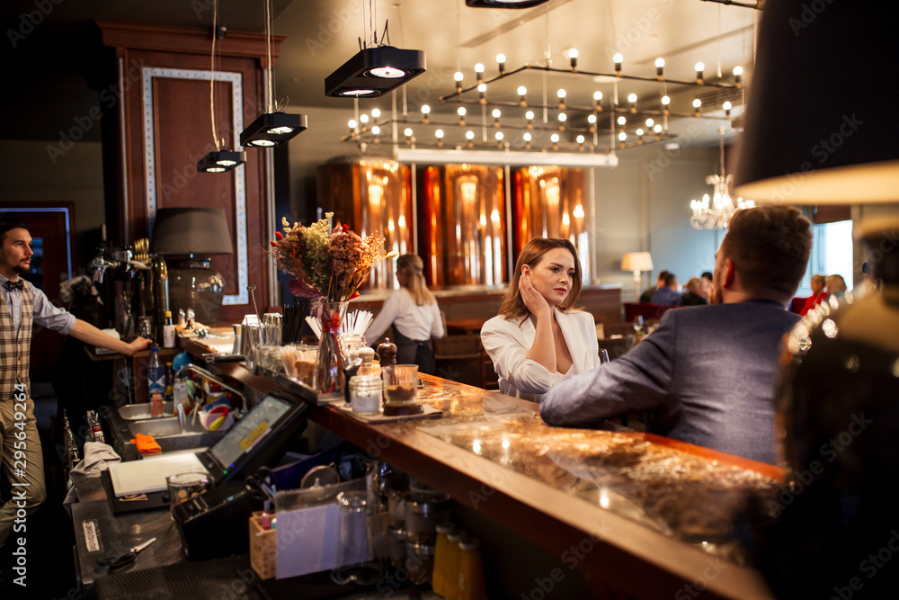 Attractive good shaped woman dressed in white clothes, touching neck thoughtfully sitting at bar counter together with handsome friend at restaurant, side view
