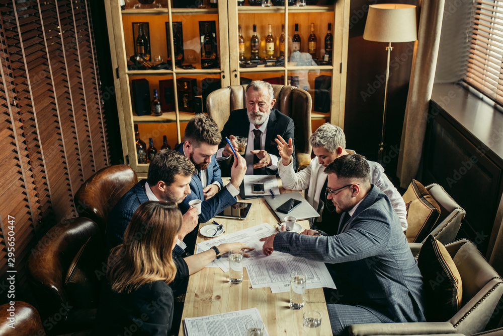 Team of specialists happily concluded important meeting. Young coworkers closed eyes with relief, raised hands in the air, Old man at the head of conference table raised a glass of whiskey